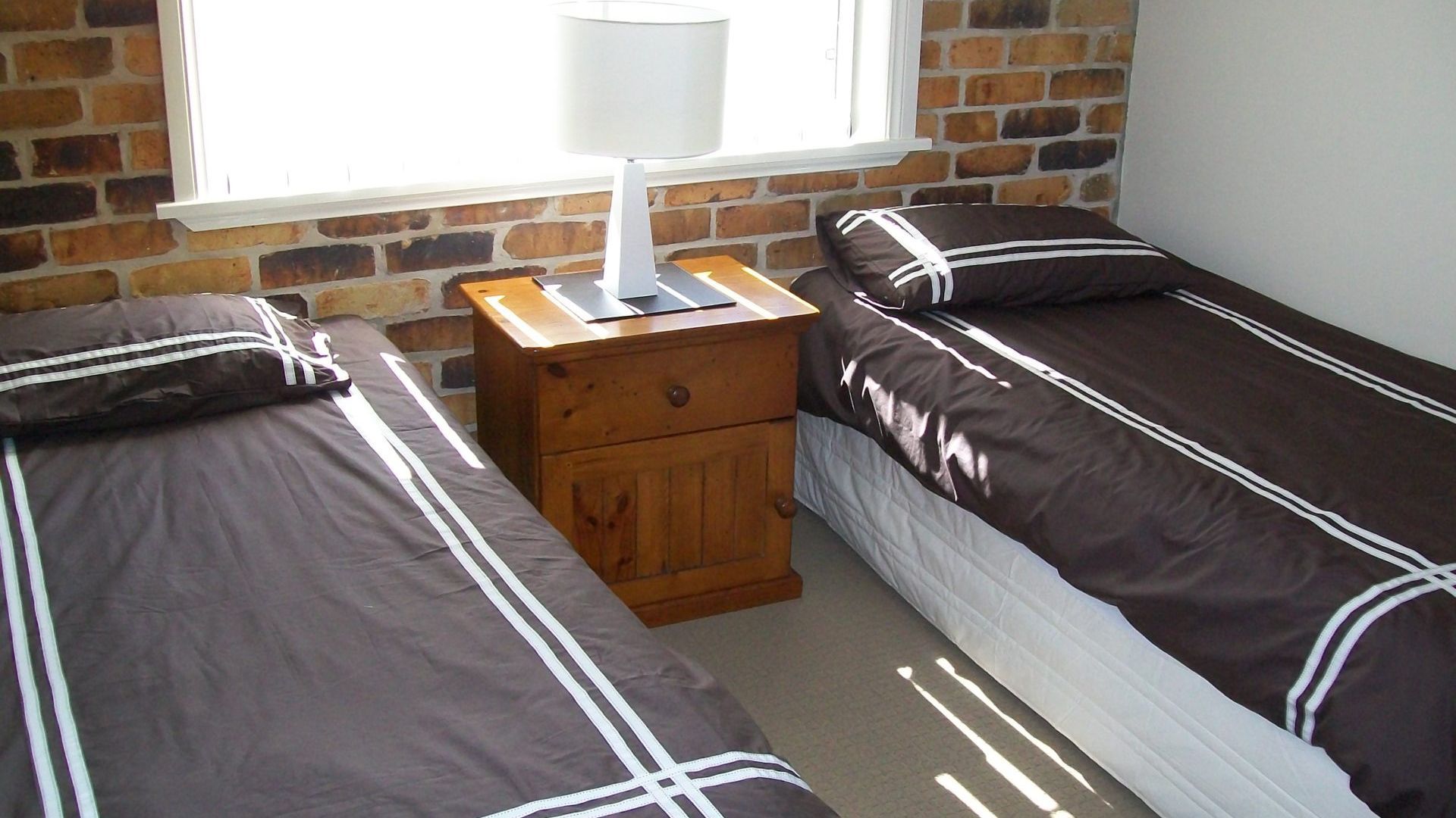 A bedroom with two single beds, a wooden nightstand, a lamp, and a brick wall.
