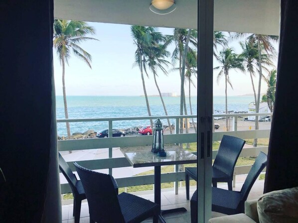 A balcony with a view of the ocean and palm trees. There is a table with chairs and a lantern.