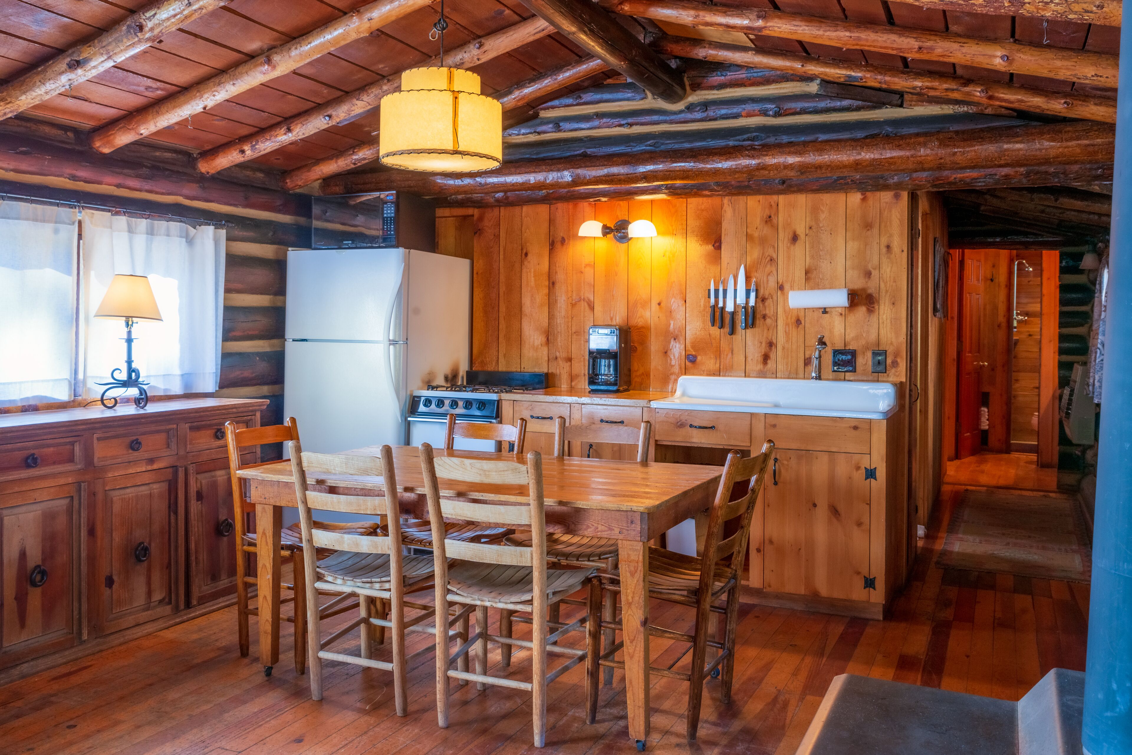 A rustic wooden cabin interior with a dining table, chairs, a refrigerator, a stove, and a sink.