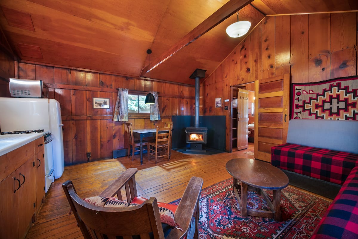 Interior acogedor de una cabaña con paredes de madera, una chimenea central, una zona de comedor y un área de estar con una manta a cuadros.