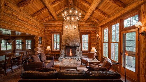 A log cabin interior with a stone fireplace, leather seating, and large windows. - Corkins Lodge (Chama)
