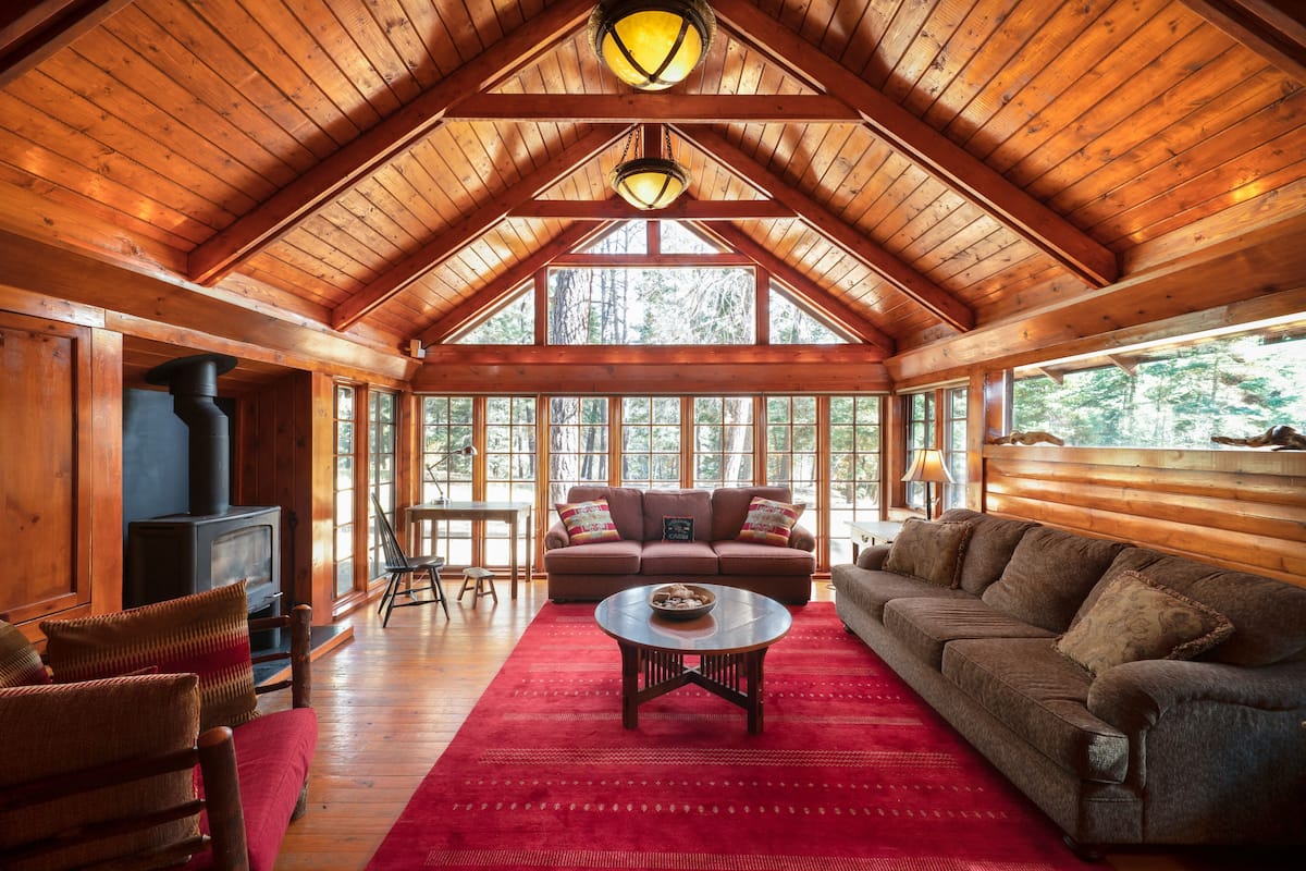 Interior acogedor de una cabaña con techo de madera, chimenea, sofá y un ventanal con vistas al bosque.