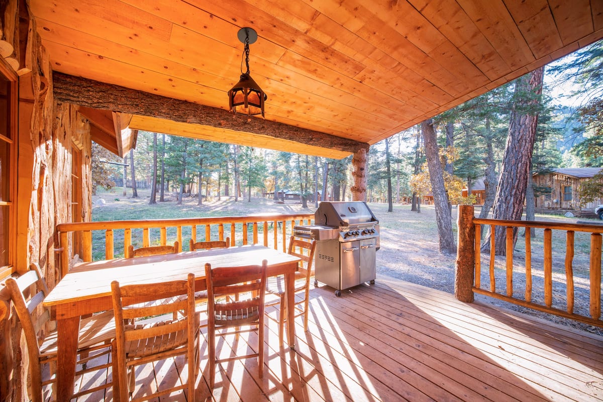 Una terraza de madera con mesa y sillas, una parrilla y vistas al bosque.