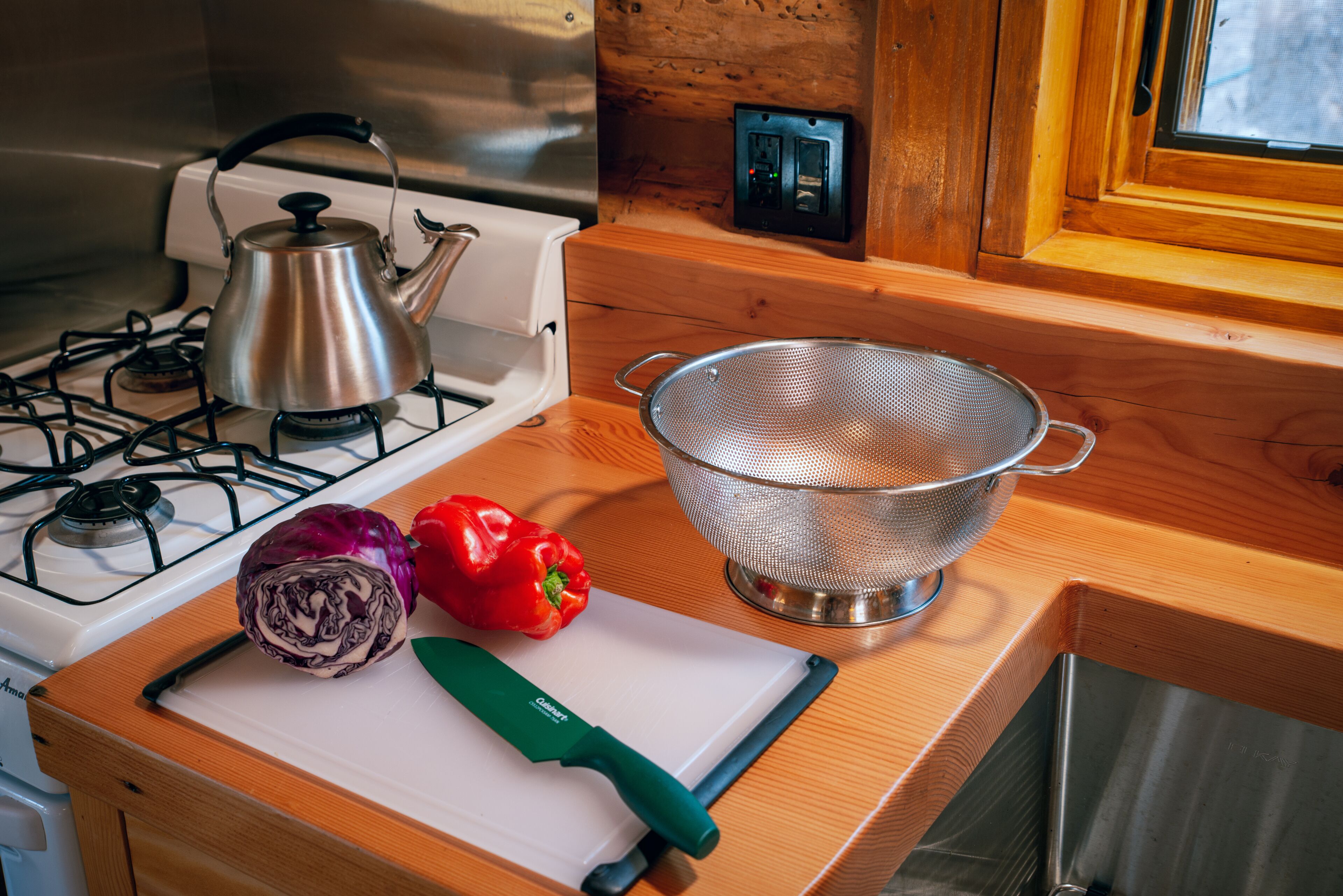 A kitchen with a gas stove, a kettle, a colander, a red bell pepper, a cabbage, and a green cutting board.