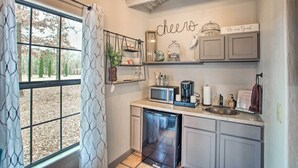 A kitchen with a built-in dishwasher, a microwave, and a coffee maker. There is a window with curtains, a shelf with decorative items, and a sign that says "Cheers Be our Guest". - ‘Blue Jay Cottage' - Pittsburg Studio on Winery! (Pittsburg)