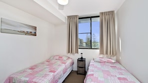 A small, clean room with two single beds, a window with curtains, a bedside table, and a wall-mounted shelf. - Sundial 503, Hollingworth Street (Port Macquarie)