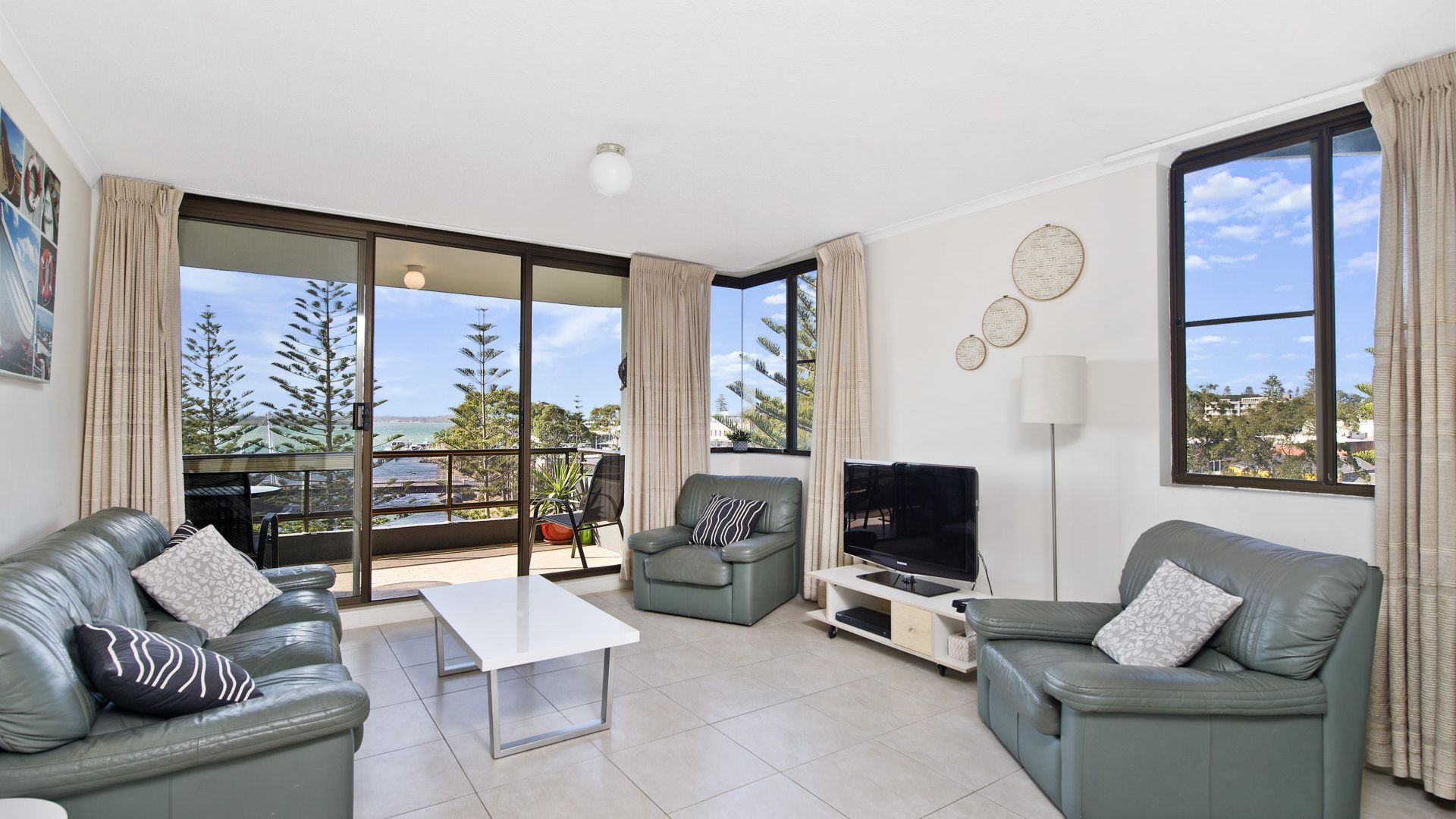 A modern living room with two sofas, a coffee table, a TV, and a balcony with a view of the sea and trees.