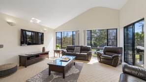 A modern living room with a flat-screen TV, leather sofas, a coffee table, and large windows offering a view of the outdoors.