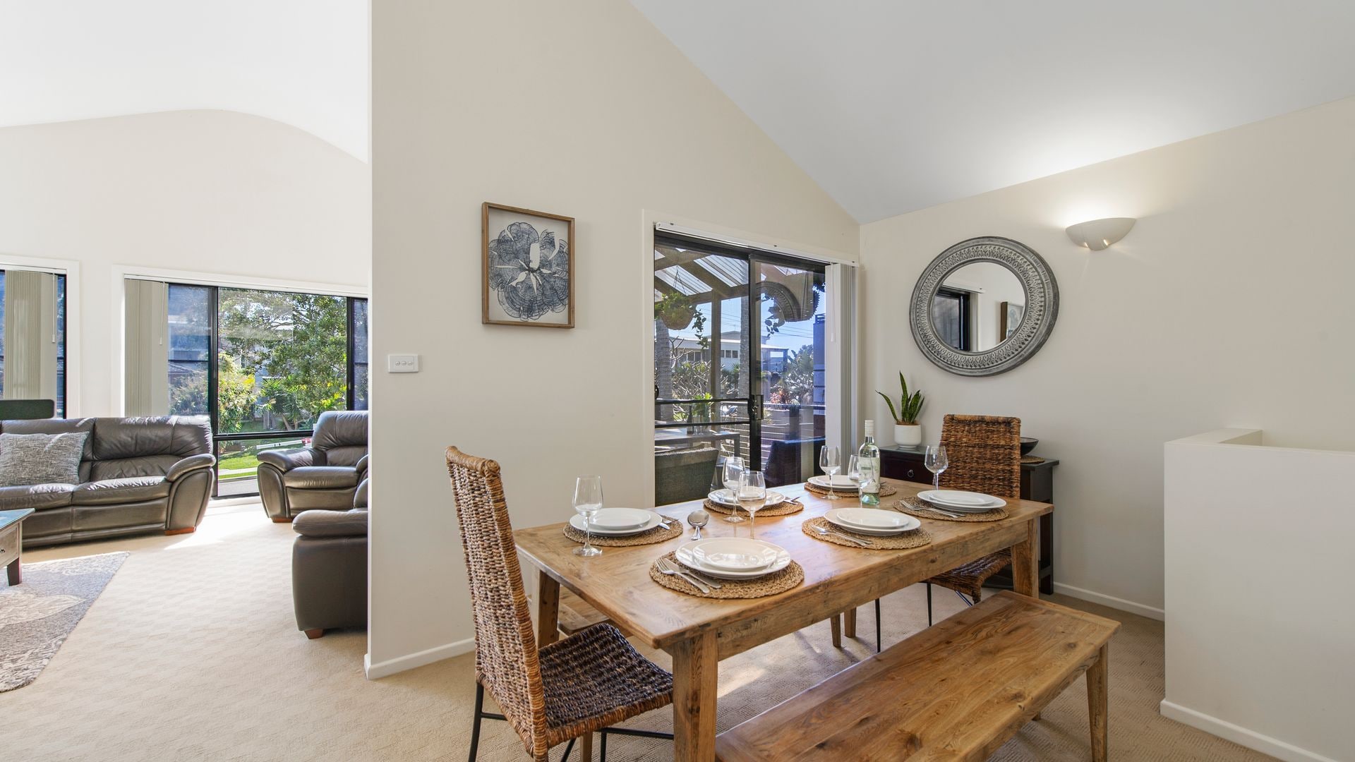 A dining area with a wooden table set for four, surrounded by wicker chairs, a mirror on the wall, and a view through a glass door to an outdoor space.
