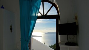 Ein Zimmer mit Meerblick, ein Fenster mit Vorhängen, ein Fernseher an der Wand und ein kleines Regal mit Gegenständen.