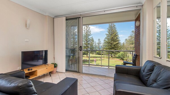 A balcony with a sliding glass door, a flat-screen TV, a wooden TV stand, a black leather sofa, and a view of a grassy area with trees. - Ocean Court 7, 13 Lord Street (Port Macquarie)