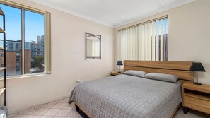 A bedroom with a bed, bedside tables, a mirror, and a window with blinds. - Ocean Court 7, 13 Lord Street (Port Macquarie)