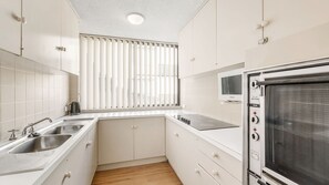 A modern kitchen with white cabinets, a stainless steel sink, a built-in oven, and a window with vertical blinds.