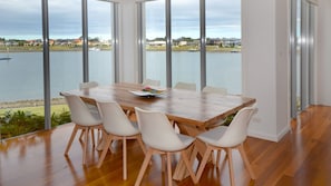 A dining area with a wooden table and white chairs, large windows with a view of a body of water and buildings. - 4/84 The Anchorage, Port Macquarie (Port Macquarie)