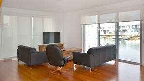 A spacious living room with a flat-screen TV, two sofas, a wooden coffee table, and a view of a canal with boats.