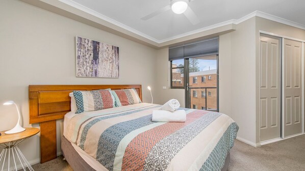 A bedroom with a bed, a bedside table, a ceiling fan, a window with blinds, and a door.