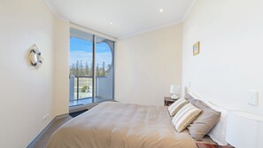 A bedroom with a large bed, a nightstand, a lamp, and a view of a balcony with trees.
