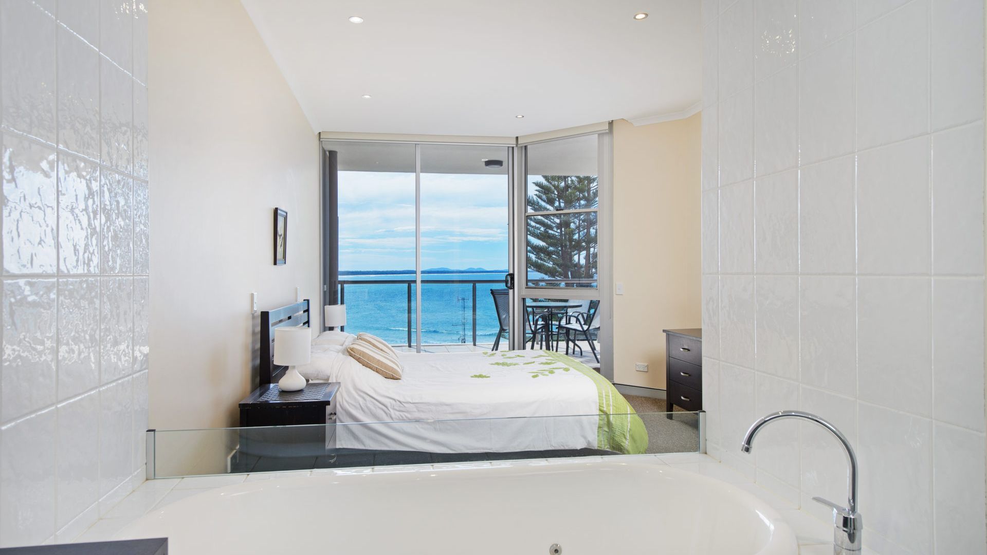A modern bedroom with a large bed, a sitting area with a view of the sea, and a separate bathroom with a bathtub.