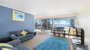 A modern living room with a blue sofa, a dining table with chairs, a large window with a view of trees and water, and a television on a stand.