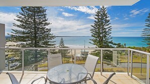 Een balkon met glazen hekwerk, een ronde tafel en twee stoelen, met uitzicht op een strand en de oceaan.