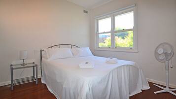 A bedroom with a bed, a bedside table with a lamp, a window, and a standing fan.