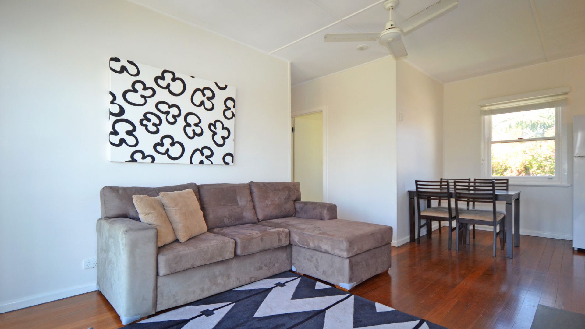 A living room with a sectional sofa, a dining table with chairs, a ceiling fan, and a window with blinds.