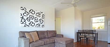A living room with a sectional sofa, a dining table with chairs, a ceiling fan, and a window with blinds.