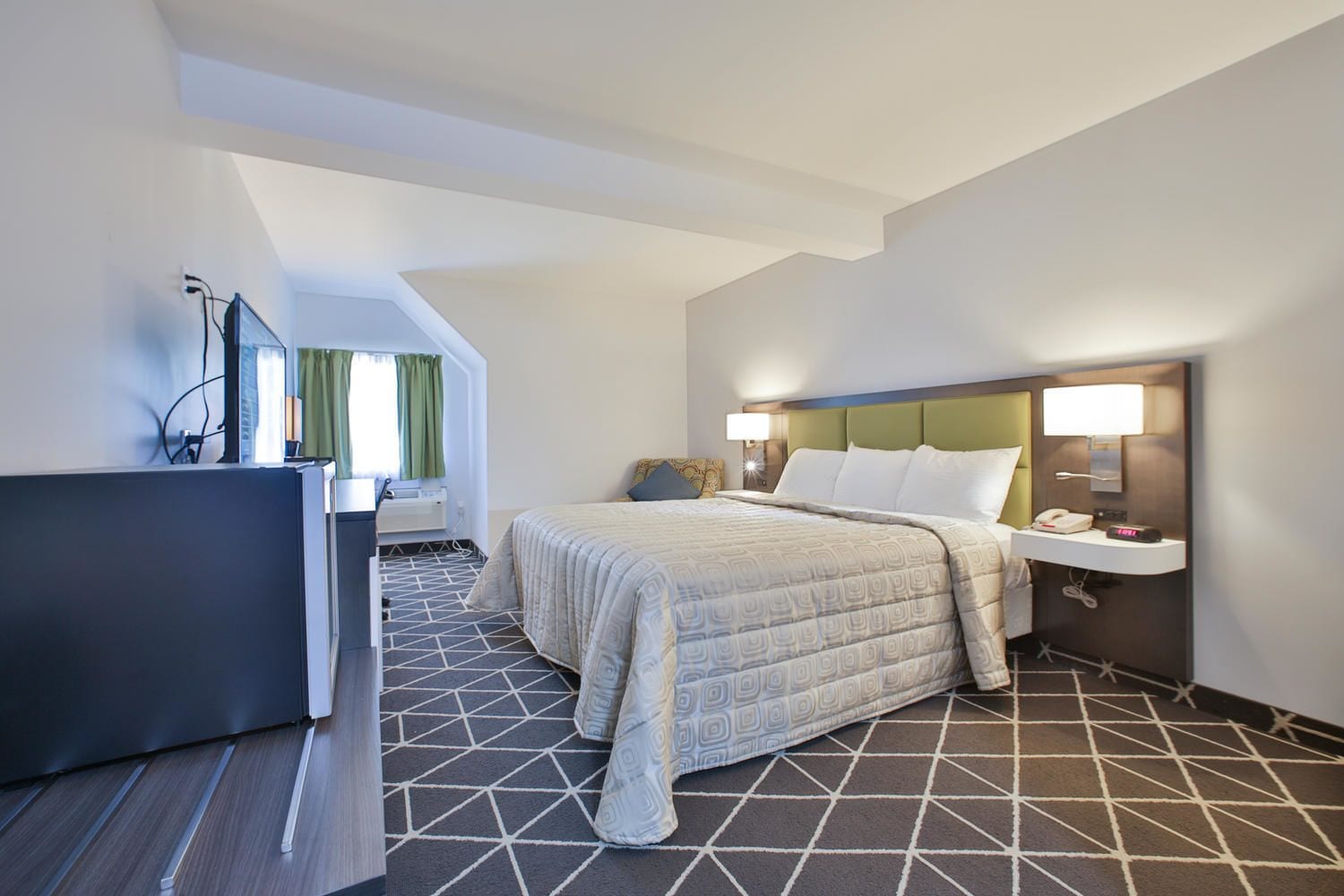 A hotel room with a bed, bedside table, lamp, and a desk with a computer.