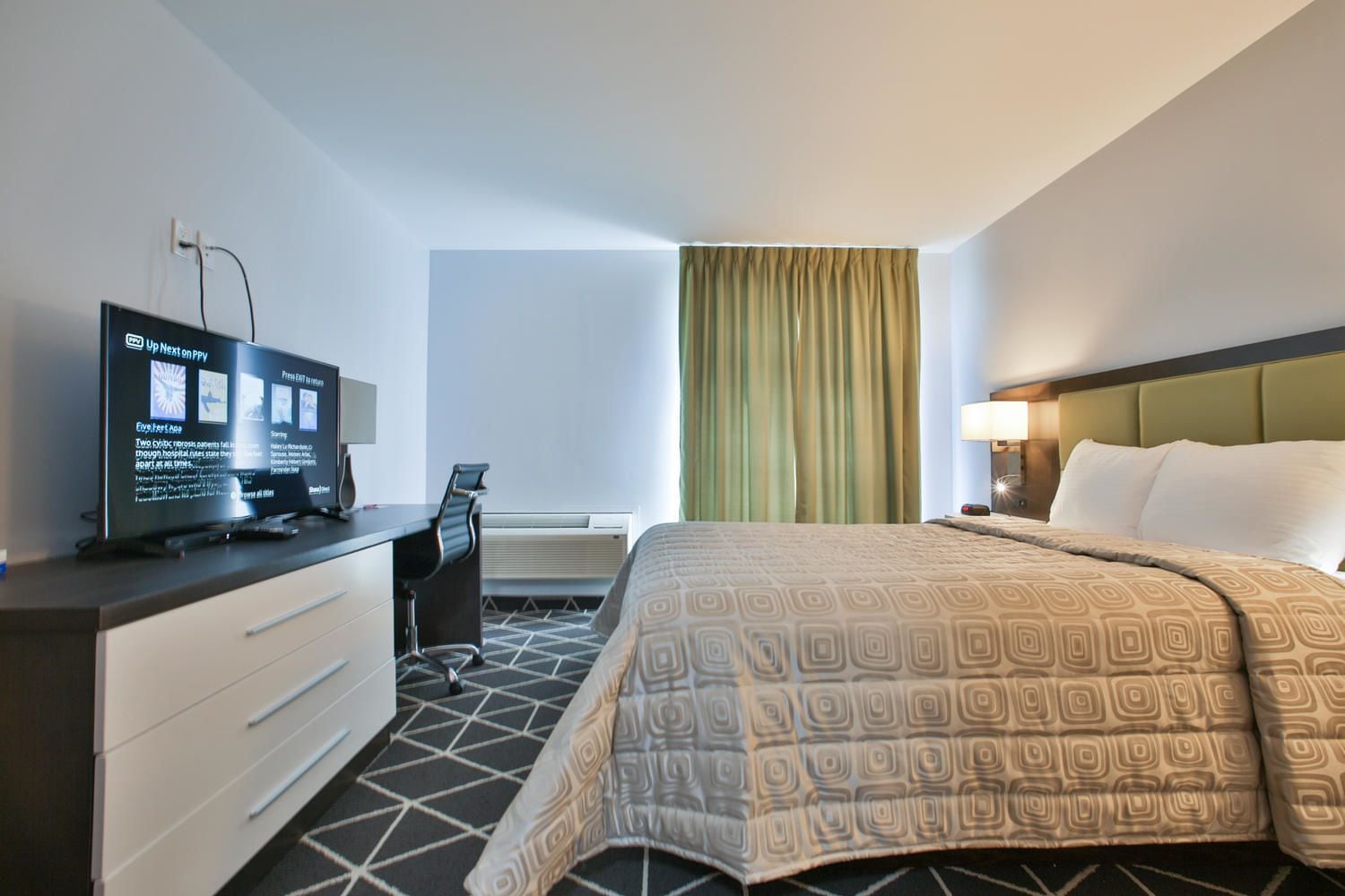 A hotel room with a bed, a desk with a computer, a chair, and a TV displaying information.
