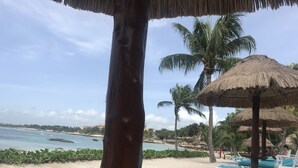 Vista da praia com guarda-sol de palha, palmeiras e cadeiras de praia.