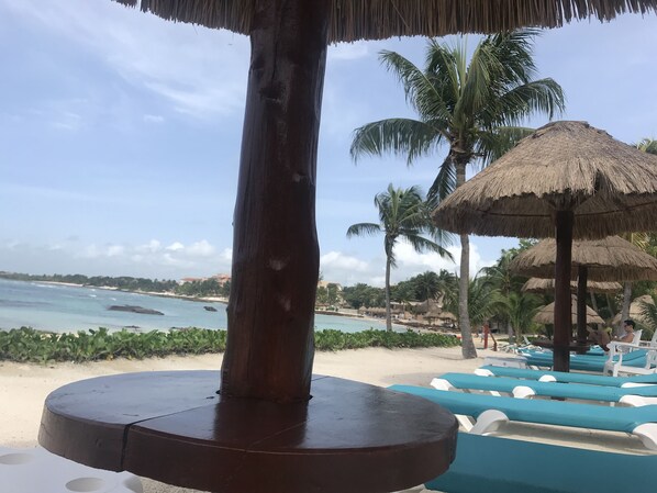 Une vue sur la plage avec un parasol en chaume, des palmiers et des chaises longues.