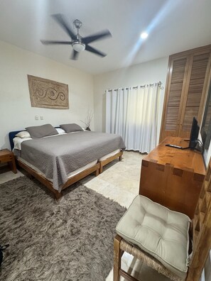 Une chambre à coucher comprenant un lit, un bureau, une chaise, un ventilateur de plafond et une fenêtre avec des rideaux.