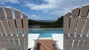 Two white Adirondack chairs facing a swimming pool with a view of the ocean and greenery.