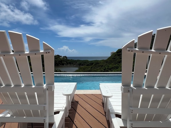 Dos sillas Adirondack blancas mirando una piscina con vistas al océano y follaje.