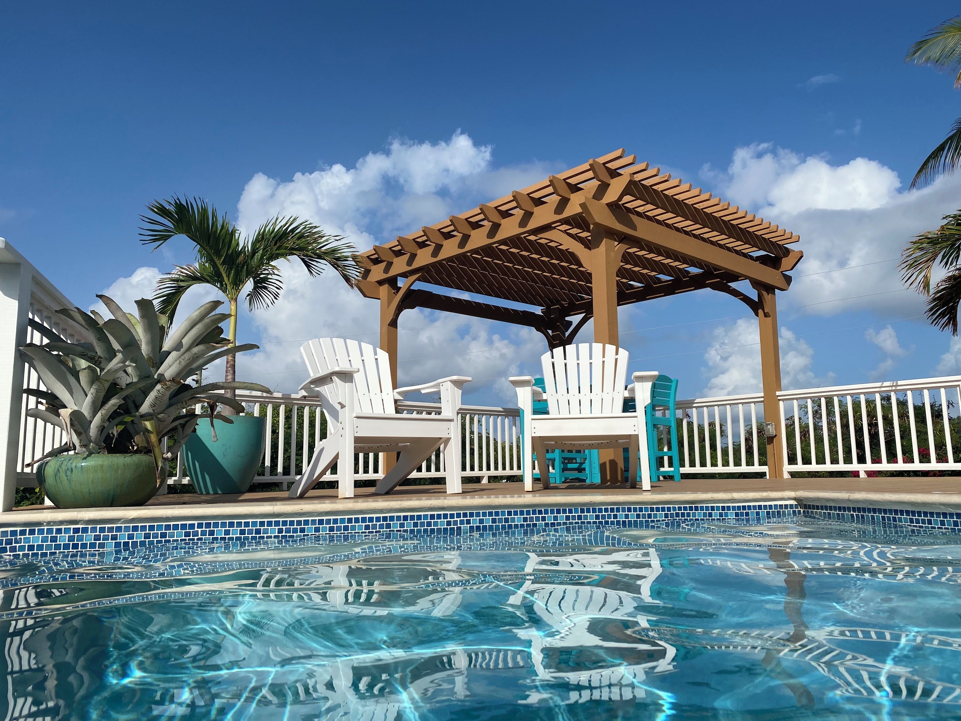 Uma área de piscina com pergolado de madeira, cadeiras Adirondack brancas e piscina revestida com azulejos.
