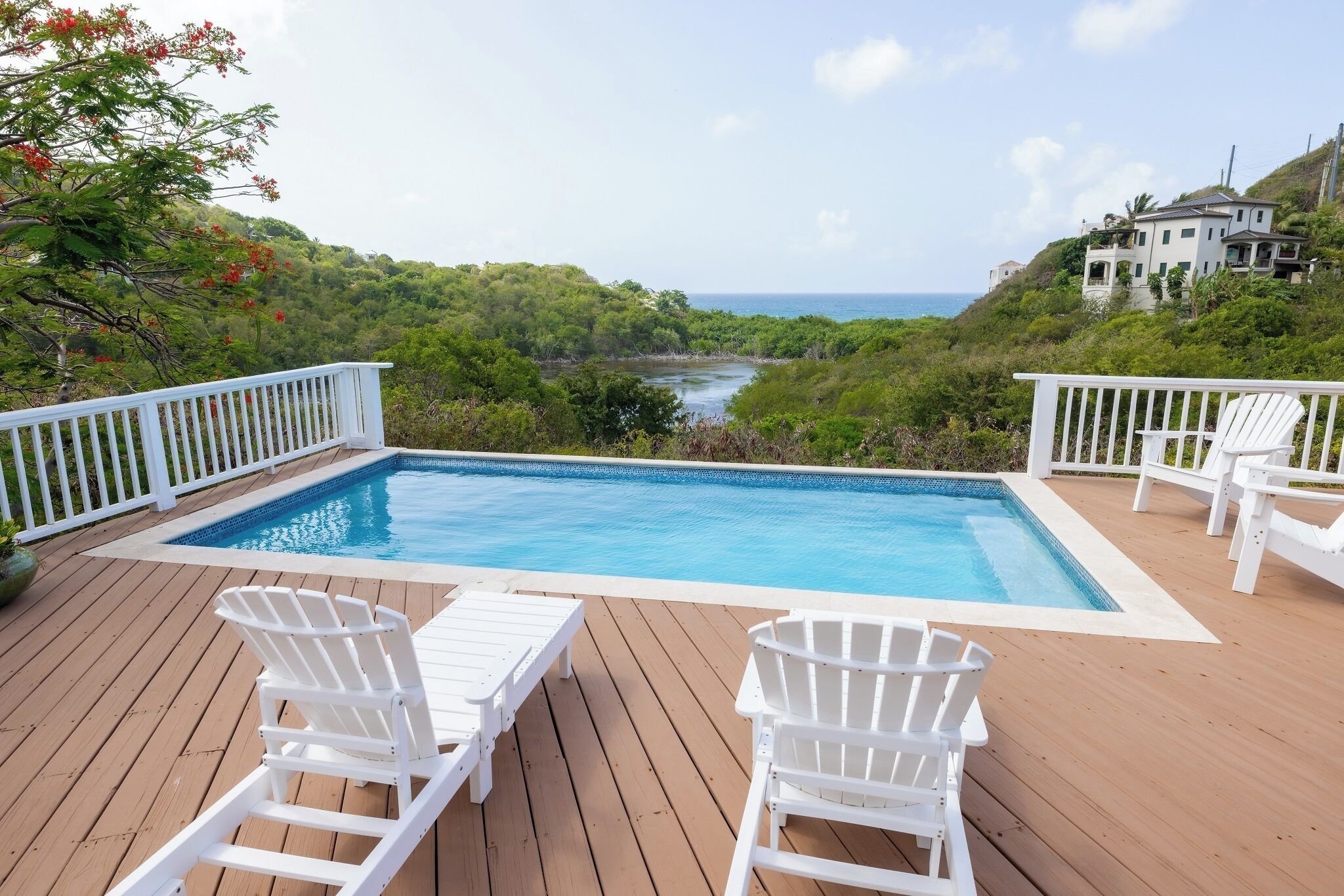 A wooden deck with a swimming pool, white deck chairs, and a view of the ocean and hills.