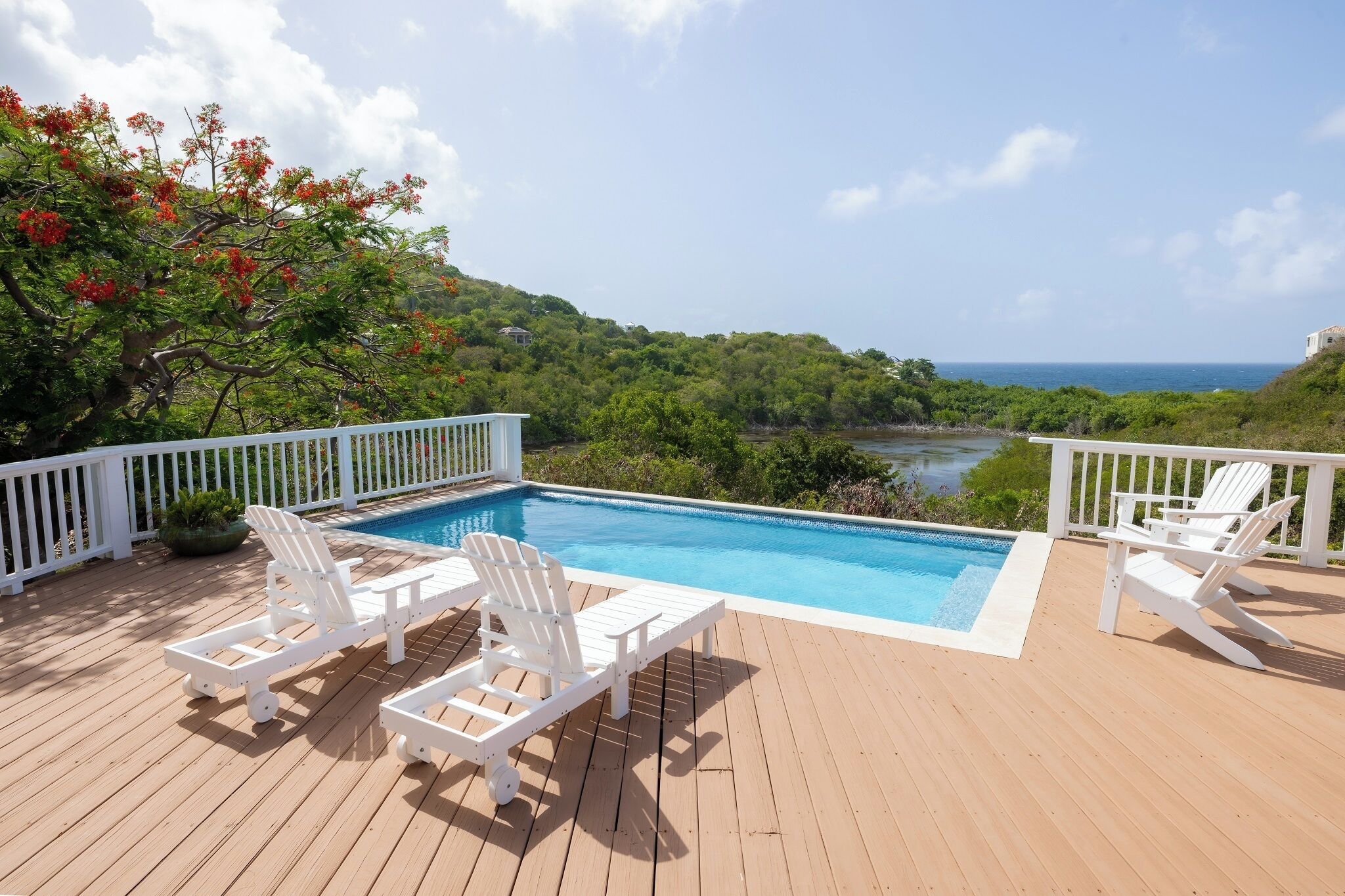 A wooden deck with a pool, white deck chairs, and a view of the ocean and greenery.