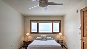A bedroom with a bed, two bedside tables with lamps, a ceiling fan, a window with blinds, and a view of the sea.