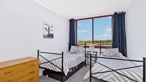 A bedroom with two beds, a wooden dresser, a window with curtains, and a cityscape view.