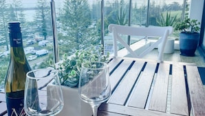 Een tafel met een fles, twee glazen, een potplant en een witte stoel, met uitzicht op een parkeerplaats en bomen.