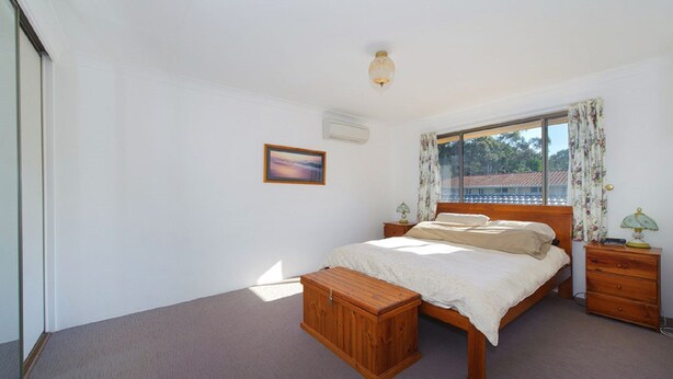 Un dormitorio con cama, mesitas de noche, un cofre de madera, una ventana con cortinas y un aire acondicionado de pared.