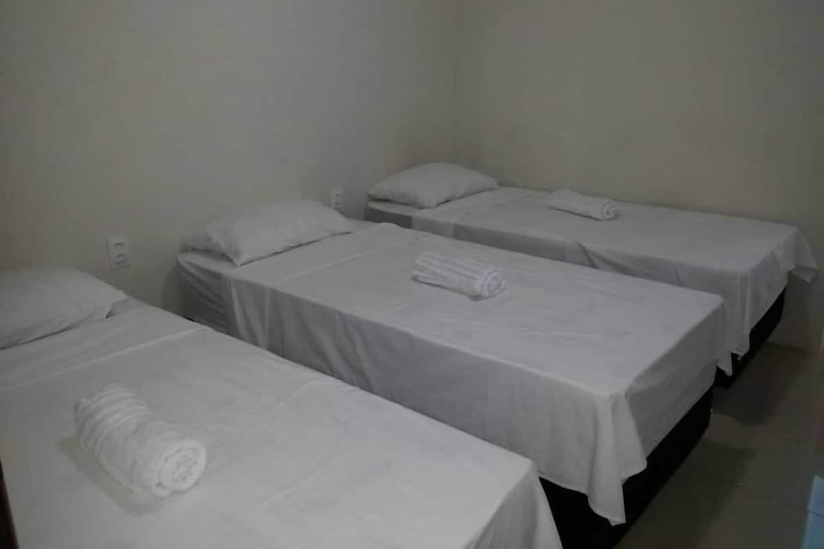 Três camas de solteiro com roupa de cama e almofadas brancas, uma parede branca e uma tomada visível.