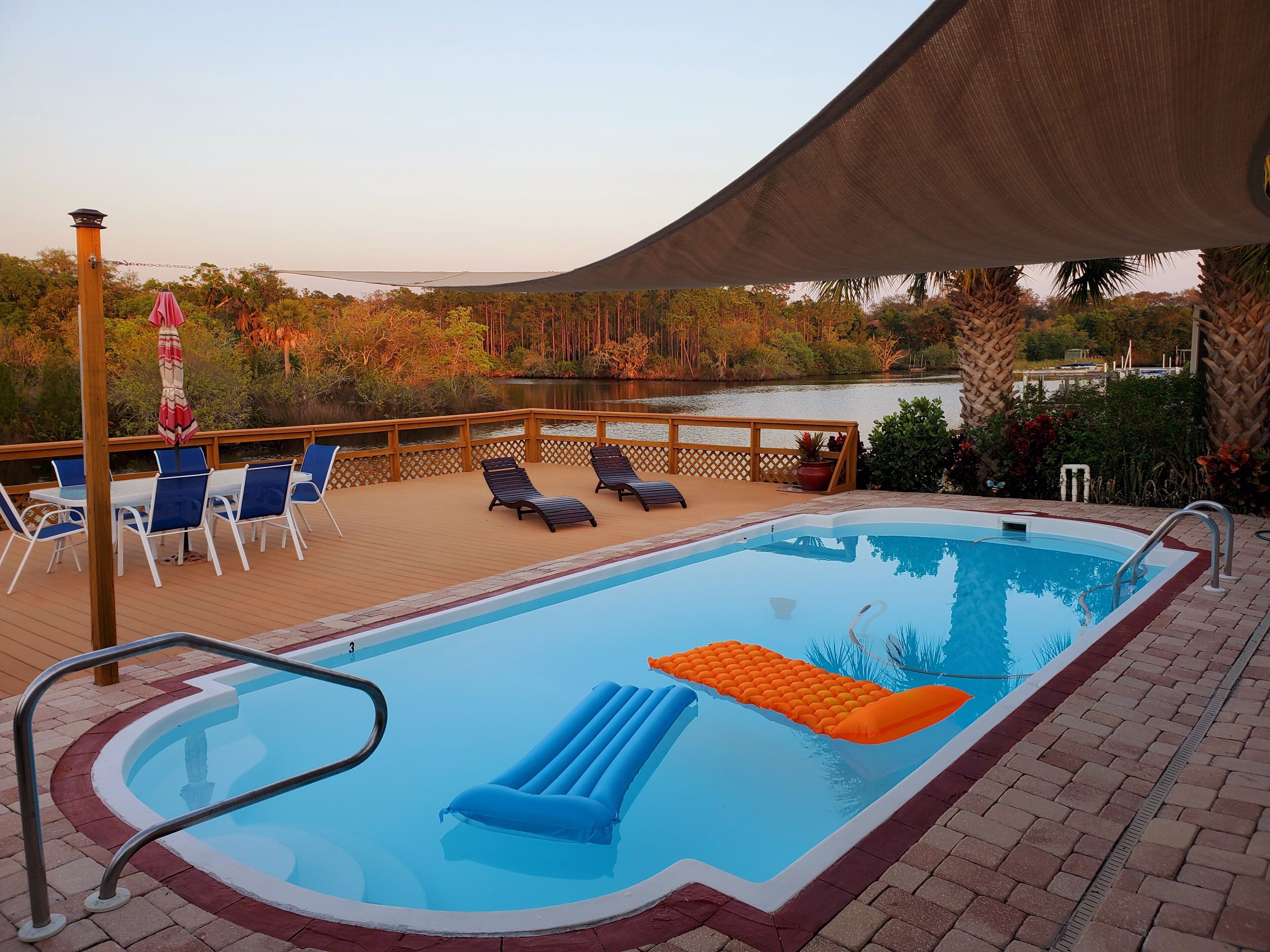 A swimming pool with lounge chairs and a table, surrounded by a wooden deck and a view of a lake.