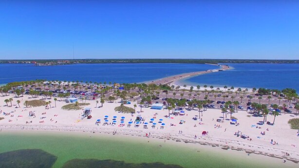 An aerial view of a beach with clear blue water, a sandy shore, and numerous umbrellas and beachgoers. - 3BR Waterfront Oasis w/ Private Solar Heated Pool and Spacious Deck (Holiday)