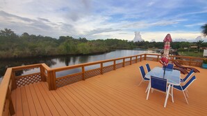 A wooden deck with a table and chairs, overlooking a calm river and lush greenery. - 3BR Waterfront Oasis w/ Private Solar Heated Pool and Spacious Deck (Holiday)