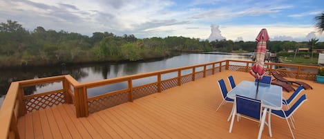 A wooden deck with a table and chairs, overlooking a calm river and lush greenery.