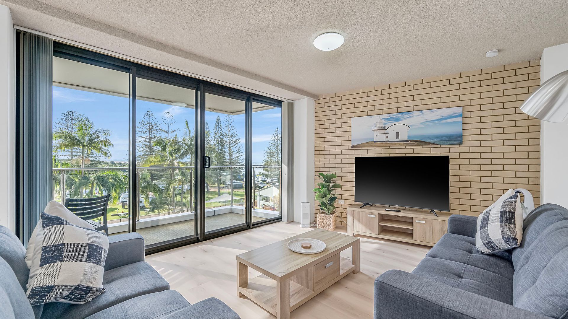 A modern living room with a flat-screen TV, grey sofas, a wooden coffee table, and a balcony with a view of palm trees and a beach.