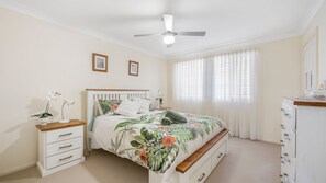 Un dormitorio con cama, mesitas de noche, cómoda, ventilador de techo y ventana con cortinas.