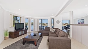 Un salon moderne avec un canapé, une table basse et un téléviseur. De grandes fenêtres offrent une vue sur l’extérieur.