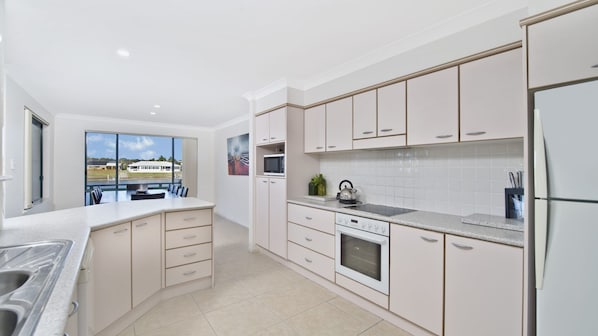 Une cuisine moderne avec des meubles blancs, un grand îlot central et une vue sur une maison par la fenêtre.